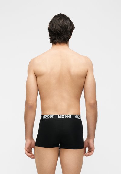 Άνδρας μοντέλο, φαίνεται από πίσω φορώντας μαύρα boxer Moschino σε απλό ανοιχτό γκρι φόντο.
