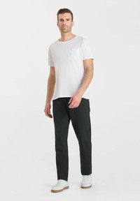 Man die staat en een wit T-shirt met korte mouwen en een borstzak draagt, zwarte broek en witte sneakers op een vrijwitte achtergrond.