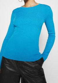 Personne portant un haut à manches longues côtelé bleu vif associé à un pantalon en cuir noir, accessoirisé d'un collier en argent et d'une boucle d'oreille en anneau.