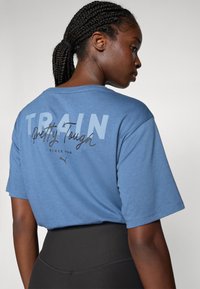 Blauwe katoenen t-shirt met korte mouwen toont vetgedrukte tekst "TRAIN" en cursieve tekst "Pretty Tough" op de achterkant. Bevat een detail van het Puma-logo.