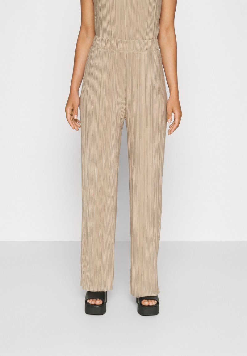 Pantalon beige plissé à jambes larges avec taille élastique porté avec des sandales noires à plateforme et orteils ouverts sur une personne debout.