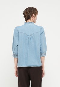 Šviesiai mėlynas džinsinis marškinis su pūstomis rankovėmis ir raukšlėtu apykakle. Yra smock'o stiliaus rankogaliai ir surinkta nugaros detalė, užtikrinanti laisvą prigludimą.