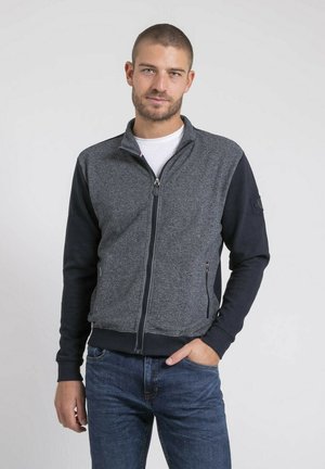 Homme aux cheveux courts et à la barbe, portant une veste zippée bicolore gris et bleu marine sur une chemise blanche, avec une main dans la poche, posant devant un fond uni.