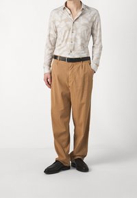 Vaaleanbeige paita, jossa on valkoinen lehtikuosi, napitettu alas, yhdistetty löysiin, vaaleanruskeisiin housuihin ja mustaan vyöhön, käytetään mustien kenkien kanssa.