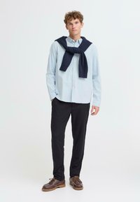 Lichtblauwe overhemdbloes gecombineerd met donkere broek, een marineblauwe trui om de nek geknoopt en bruine veterschoenen. Eenvoudig, casual ontwerp.