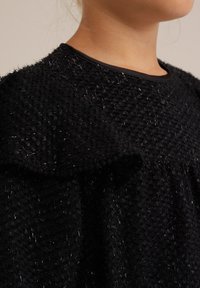 Črn top iz teksturirane tkanine, ki ima puhasto, neenakomerno površino in sijoče kovinske niti, s gladkim črnim ovratnikom in nežnim nabranim detajlom.