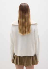 Cremfarbener Strickpullover mit geripptem Kragen und Saum, kombiniert mit einem grünen Cordrock. Lange glatte Haare sind von hinten sichtbar.