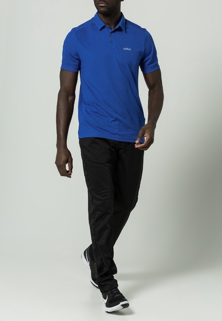 Homme marchant portant un polo à manches courtes bleu, un pantalon noir et des chaussures de sport noires et blanches sur un fond clair uni.