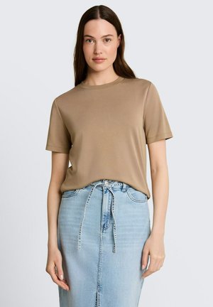 Frau mit langen braunen Haaren, die ein beiges Kurzarmshirt trägt, das in einen hellblauen Jeansrock mit vorderem Bindegürtel gesteckt ist, steht vor einem grauen Hintergrund.