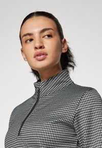 Fekete-fehér mintás cipzáras felső magas gallérral és texturált anyaggal, keskeny fazonnal és finom fényes hatással.