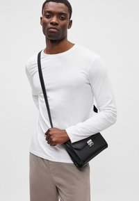 Juodas odinis crossbody krepšys su sidabro spalvos detalėmis, lygia tekstūra ir uždarymu su atvartu. Modelis dėvi baltą ilgomis rankovėmis marškinį ir smėlio spalvos kelnes.