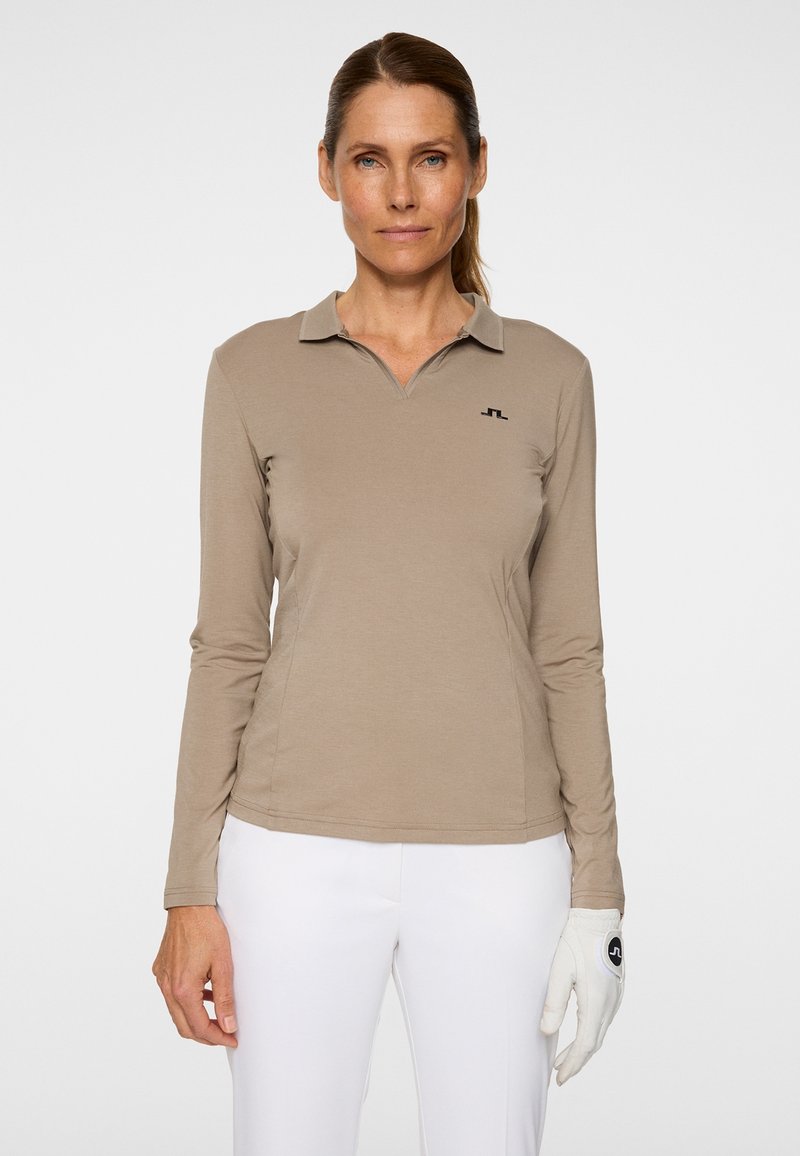 Hellbraunes Langarm-Poloshirt mit Kragen, das ein kleines schwarzes Logo auf der Brust zeigt, kombiniert mit weißen Hosen und einem weißen Handschuh.