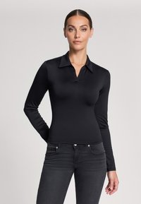 Maglietta polo nera a maniche lunghe con colletto a V, realizzata in tessuto liscio. Abbinata a jeans scuri aderenti. Design semplice, senza pattern o decorazioni visibili.