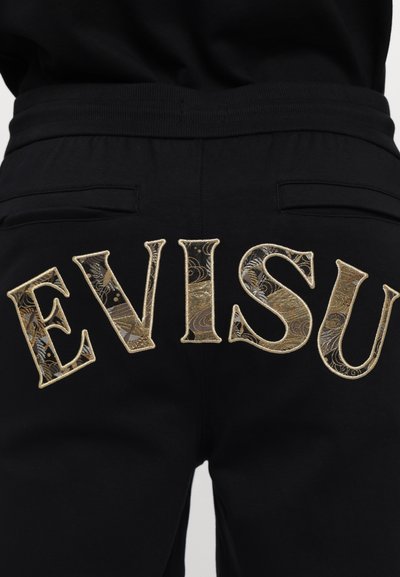 Jogging en noir avec de grandes lettres dorées brodées "EVISU" au dos, ornées de détails complexes dans la broderie et deux poches latérales.