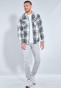 Schwarz-weiß kariertes Hemd mit zwei Brusttaschen, langen Ärmeln und Reißverschluss vorne, kombiniert mit hellgrauen Jeans und weißen Turnschuhen.