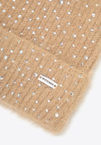 Béžová pletená tkanina s žebrovanou texturou, zdobená rozptýlenými stříbrnými třpytkami. Obsahuje malou kovovou cedulku s nápisem "WITTCHEN".