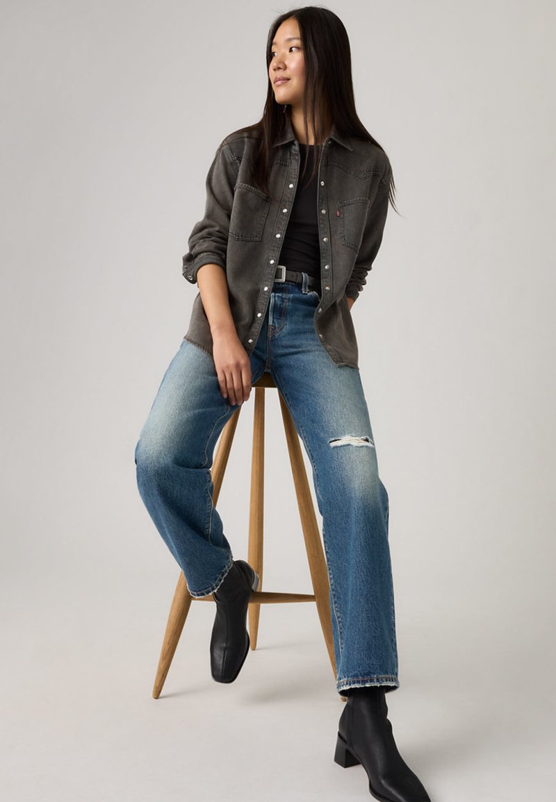 Woman with long dark hair wearing a black shirt, gray button-up jacket, ripped blue jeans, and black boots sits on wooden stool against gray background.
