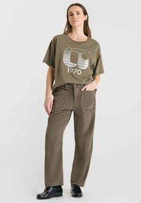 Olijfgroene katoenen t-shirt met een grote witte graphic en de tekst "1970", gedragen met bruine corduroy broek en zwarte schoenen.