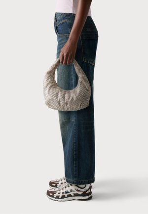 Sac à main tissé beige avec une silhouette arrondie, surface texturée et une large bandoulière, associé à un jean bleu taille haute et des baskets marron.