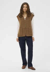 Smešani smeđi vuneni prsluk sa patentnim zatvaračem, dvostrukim proširenim džepovima i visokim izrezom, uparen sa tamnoplavim farmerkama i smešanim smešanim cipelama.