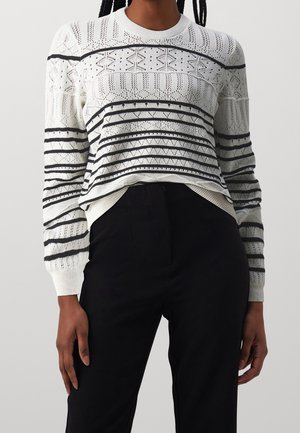 Personne portant un pull en maille blanc avec des rayures géométriques noires et un pantalon noir taille haute, debout devant un fond uni.