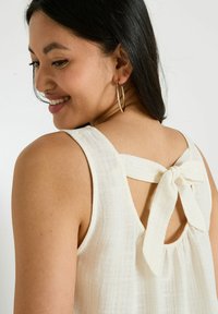 Débardeur blanc texturé avec un dos en trou de serrure et un détail de nœud. Présente des bretelles larges et une coupe décontractée. Boucles d'oreilles dorées visibles.