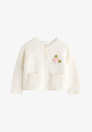 Krémový pletený detský kardigán so zapínaním na gombíky vpredu, dvoma malými vreckami a vyšívanou ružovou kvetinou so zelenými listami na hrudi.