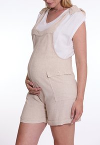 Beige linnen zwangerschapsoverall met een shorts ontwerp, voorzien van zijzakken, schouderbandjes en een relaxte pasvorm, gedragen over een wit shirt.