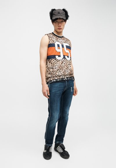 Jeune homme portant un haut sans manches imprimé léopard avec le numéro 95, un jean bleu, des baskets noires et une casquette avec des rabats d'oreilles en fourrure, debout devant un fond blanc.