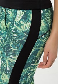 Leggings con un estampado de hojas tropicales verdes, con paneles laterales negros y acentos cosidos, que muestran una tela elástica y texturizada.