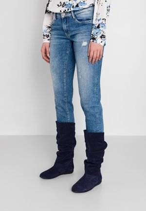 Person wearing faded blue jeans with slight distressing, floral long-sleeve top, and slouchy navy blue mid-calf boots standing on gray floor.