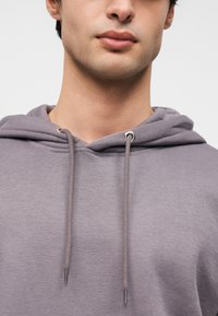 Γκρι φούτερ με μαλακή υφή, με κουκούλα με κορδόνι και μεταλλικά υφάσματα. Στενή εφαρμογή που αναδεικνύει το λαιμό και το πρόσωπο.