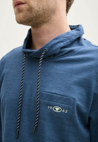 Niebieska bluza z kapturem, wykonana z teksturowanej tkaniny, z kieszenią kangurką i czarno-białymi sznurkami. Logotypy umieszczone na klatce piersiowej.