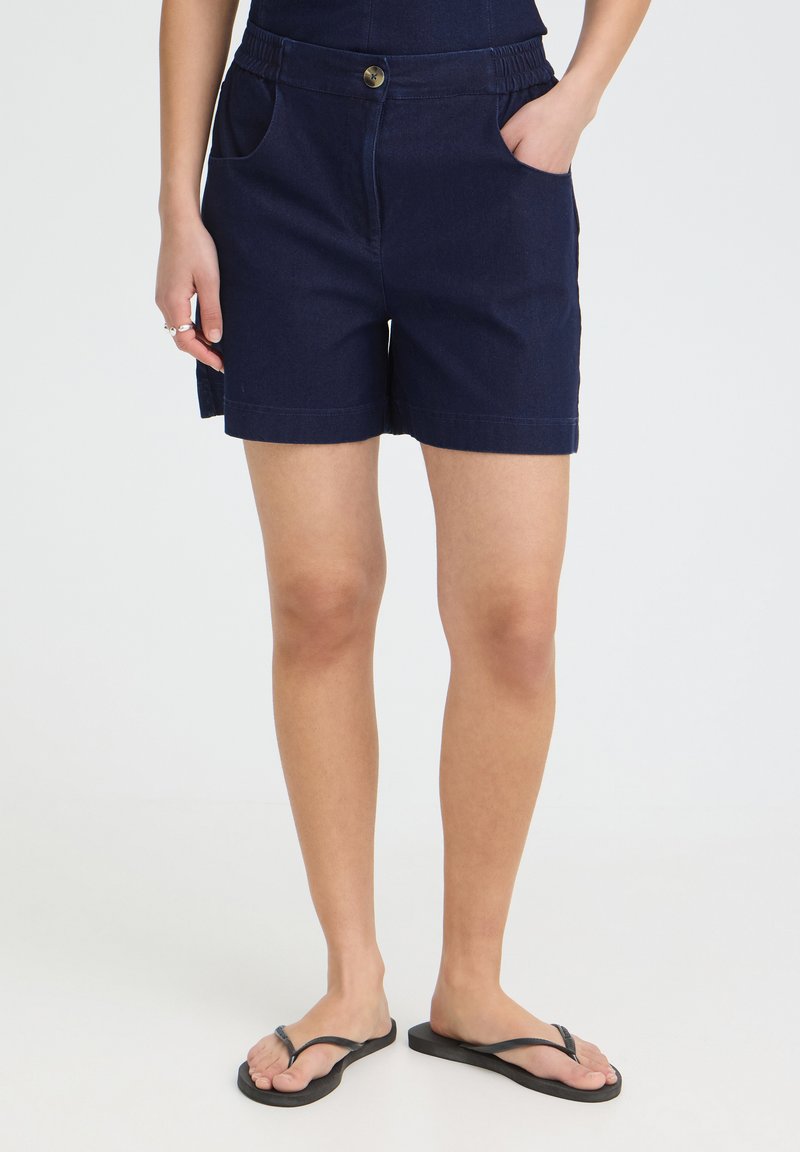 Femme portant un short bleu marine avec des poches et des tongs noires, debout devant un fond clair uni.