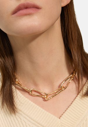 Collana di catena d'oro con maglie allungate e texture, aderente attorno al collo, abbinata a un maglione beige chiaro.