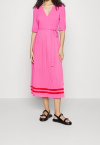 Rosa Faltenkleid mit Wickeldesign, kurzen Ärmeln und einem Band in der Taille, mit roten Streifen am Saum. Getragen mit schwarzen Sandalen.