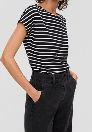 Žena nosící černobíle horizontálně pruhované tričko s krátkými rukávy zastrčené do černých džín s vysokým pasem, ruce v kapsách.