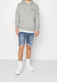 Šedá mikina s kapucí s modrým logem, džínové šortky s ohrnutými lemy a bílé vysoké tenisky. Na šortkách jsou patrné viditelné opotřebení.