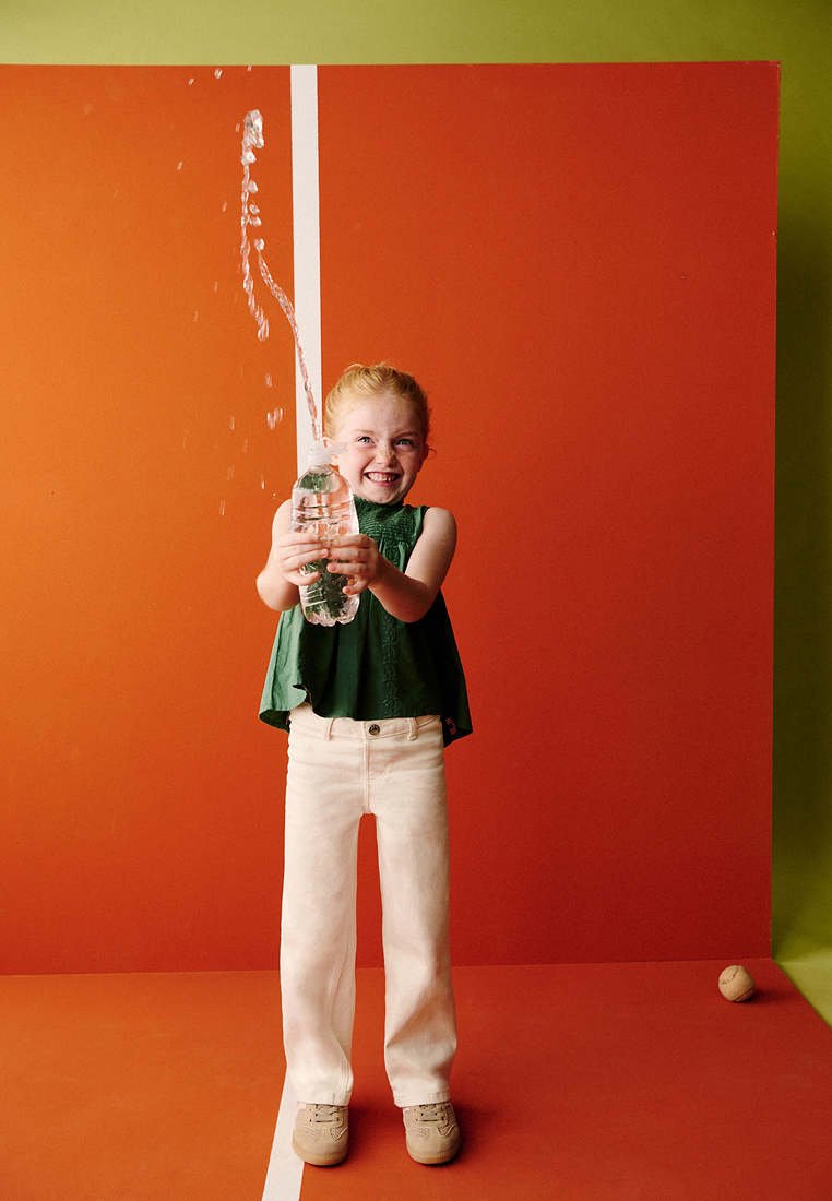 Jeune fille souriante tenant une bouteille d'eau qui projette de l'eau vers le haut, debout sur un sol orange avec un mur de fond orange et vert.