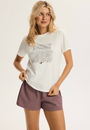 Blonde Frau trägt weißes T-Shirt mit Skizze eines Konditoreiladens und roten karrierten Shorts und steht vor schlichtem Hintergrund.
