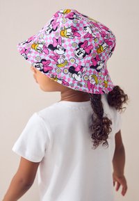 Chapeau bob rose avec un motif cartoon comprenant des fleurs et des personnages. Le matériau semble léger, avec une texture douce. T-shirt blanc visible.