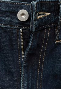 Tmavě modré džíny z denimu mají kovový knoflík s vyraženým rokem "1984", zipový zavírání a béžové prošívání podél švů.