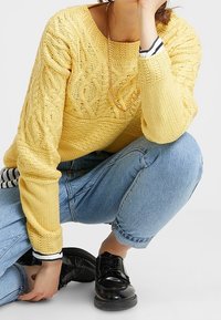 Žlutý pletený svetr s propracovanými copánkovými vzory, doplněný o zastrčené světle modré džíny a černé lakované boty.