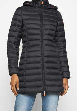 Kobieta w dopasowanej czarnej pikowanej kurtce z kapturem, zapinanej z przodu na zamek, w połączeniu z niebieskimi dżinsami, stojąca na tle jednolitego tła.