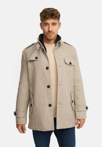 Manteau beige en mélange de laine avec des boutons noirs, des épaulettes et un col montant. Comprend des poches latérales et une coupe décontractée. Porté par-dessus un pull crème.