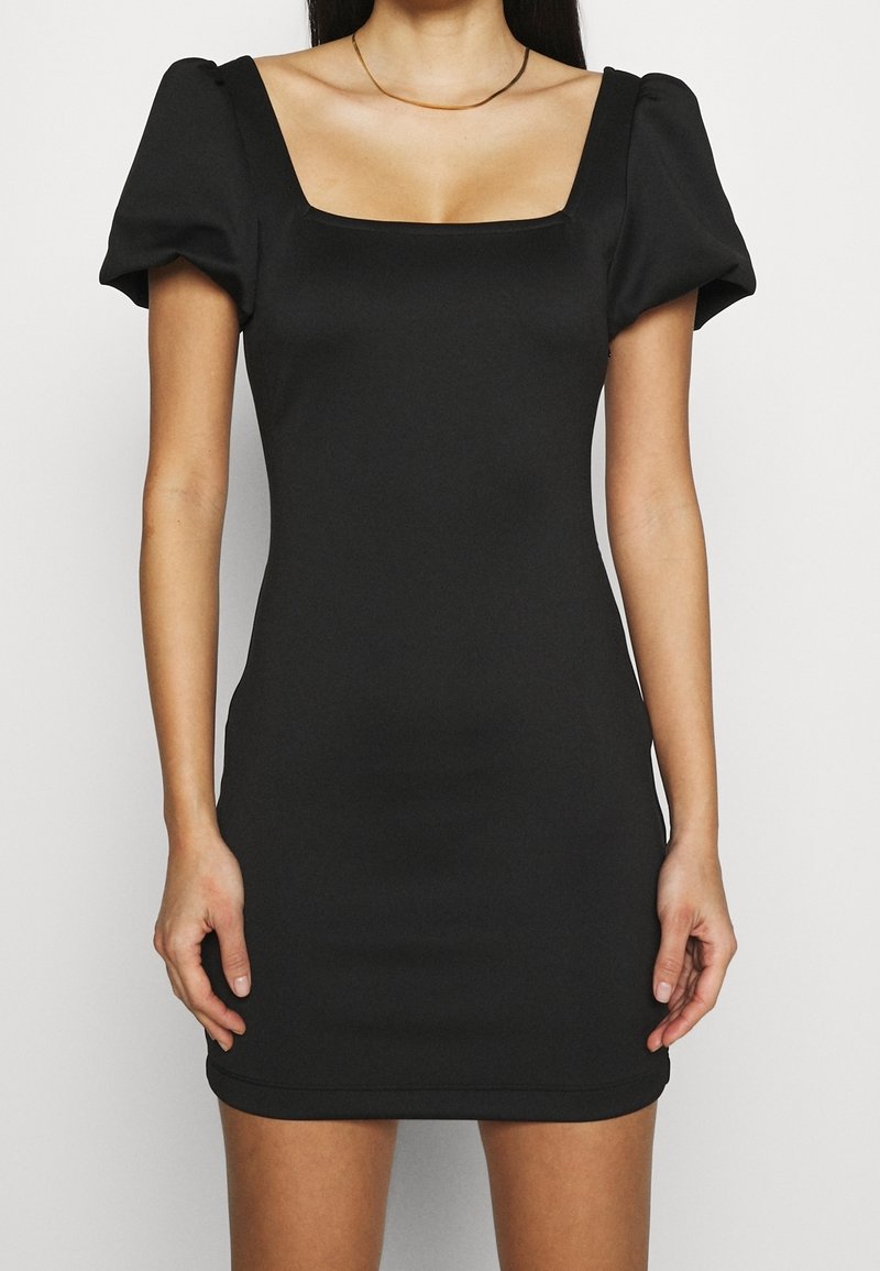 Femme portant une robe noire ajustée avec des manches courtes bouffantes et un décolleté carré, debout devant un fond clair uni.
