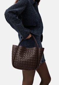 Borsa tote in pelle intrecciata marrone scuro con parte superiore arrotondata e manici robusti, esposta su uno sfondo di tessuto blu scuro texturizzato.