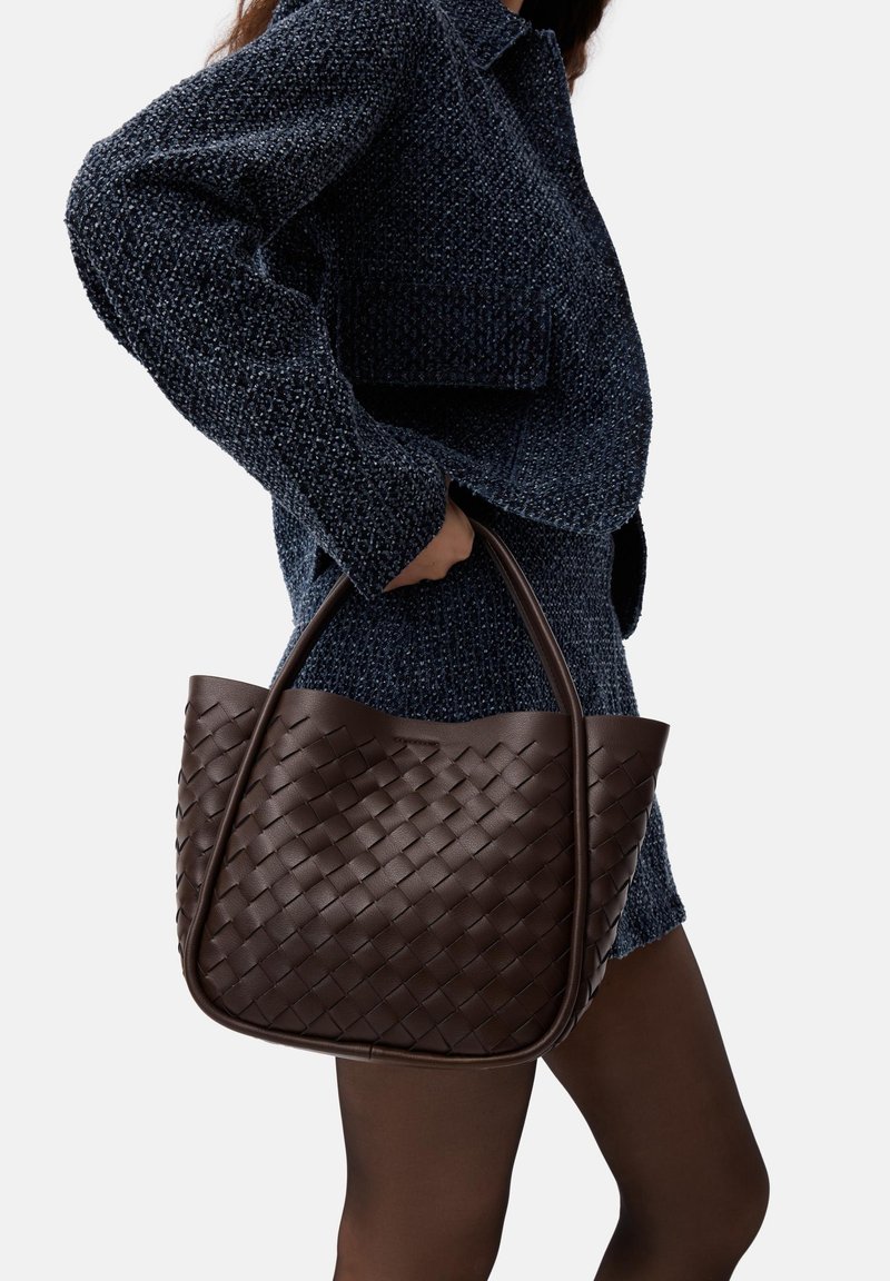Borsa tote in pelle intrecciata marrone scuro con parte superiore arrotondata e manici robusti, esposta su uno sfondo di tessuto blu scuro texturizzato.