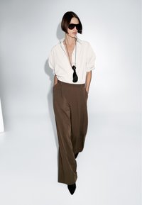 Vrouw met kort haar die grote zwarte zonnebrillen draagt, een wit overhemd met knopen, een paarse brede broek en zwarte puntige schoenen, poseert in een studio.