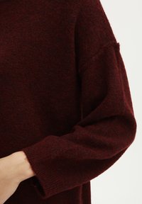 Maglione lavorato a maglia bordeaux con finitura testurizzata, caratterizzato da una vestibilità ampia e maniche larghe. Il polsino presenta un sottile motivo a costine.